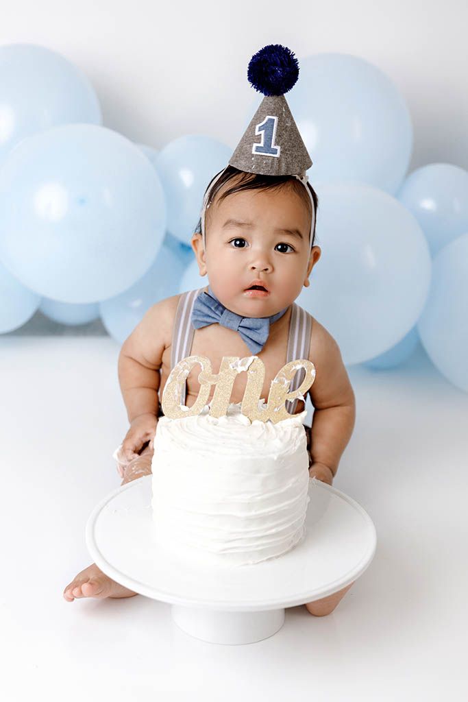 Baby boy in party hat and bow tie sits by 1st birthday cake, surrounded by blue balloons by French Valley baby photographer.