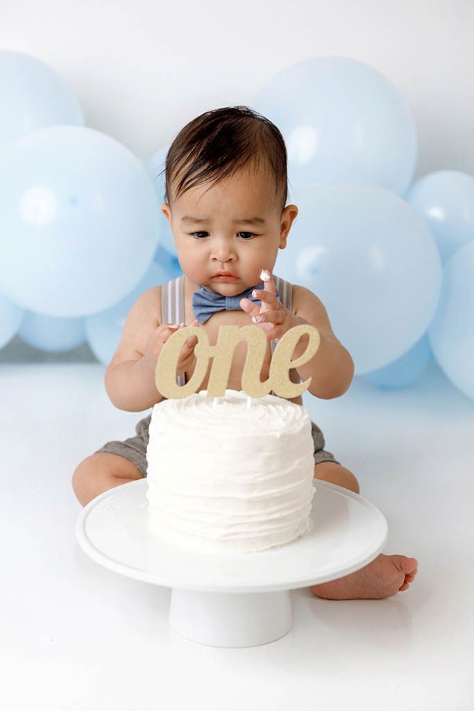 Curious baby boy in bow tie reaches toward white first birthday cake with gold 'one' topper and blue balloons behind.