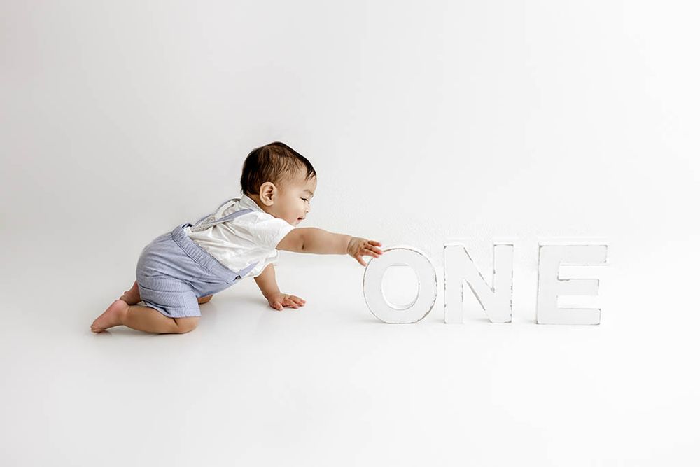 Affordable baby photographer near French Valley CA - baby boy in blue romper reaching for large white ONE letters on clean white background.