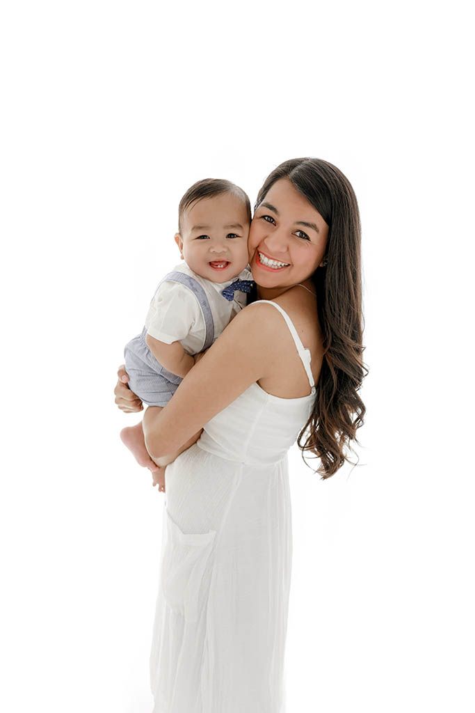 Smiling mom in white dress holding a happy baby boy against white backdrop by affordable French Valley baby photographer.