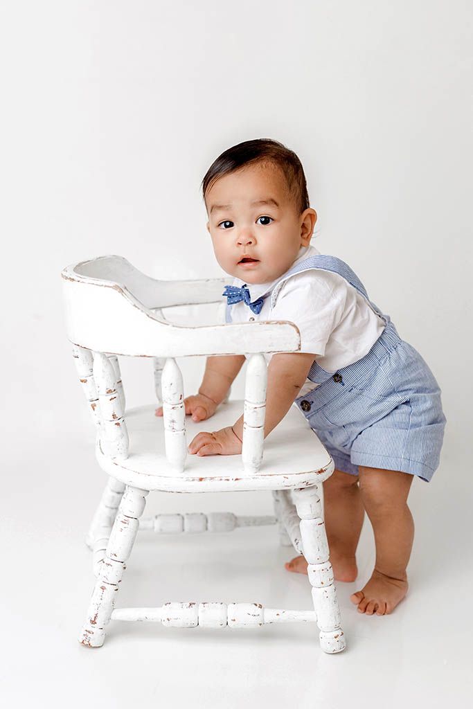 French Valley baby photographer photo of baby boy in blue suspender shorts and bow tie stands holding a vintage wooden chair.