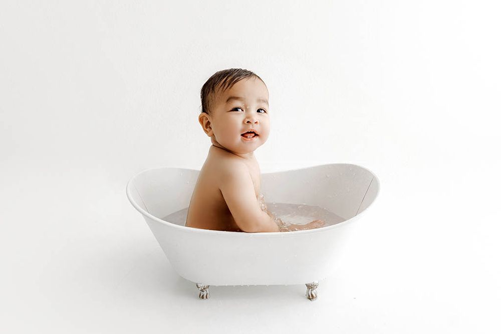 A smiling baby sits in a small white clawfoot bathtub against a white background, by French Valley baby photographer.