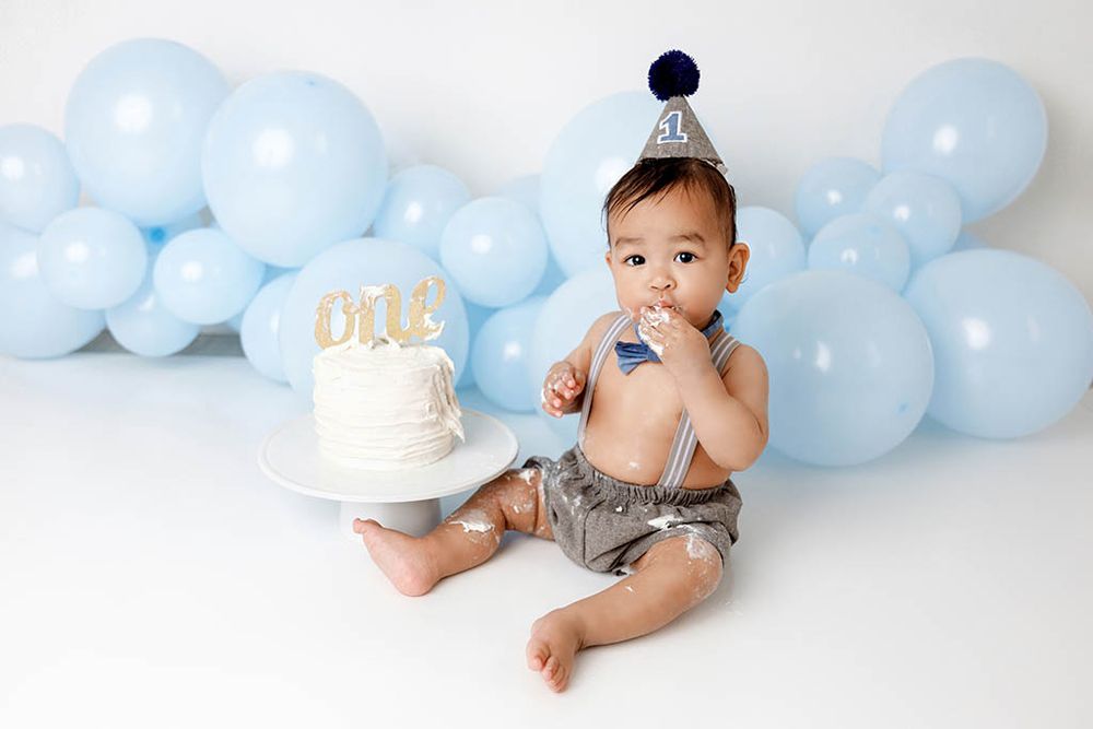 Baby boy in party hat sits with white cake and blue balloons at first birthday cake smash photo session by French Valley baby photographer.