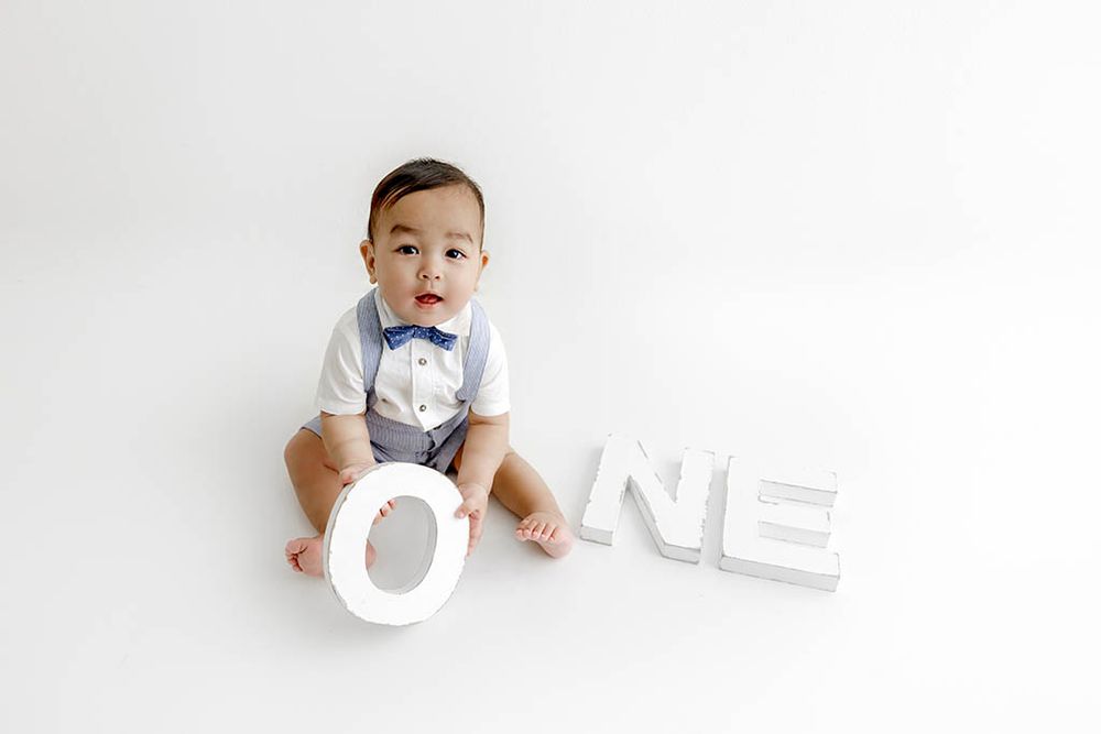 Baby boy in suspenders and bow tie sits beside large white ONE letters for first birthday photo by French Valley baby photographer.
