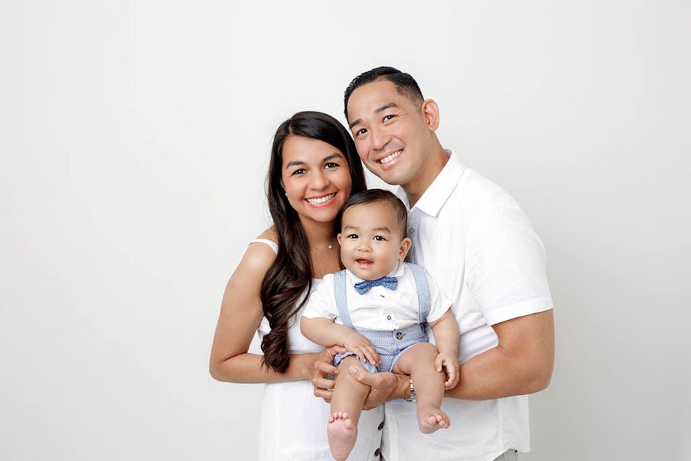 Happy young family with smiling parents holding their infant son in a blue bow tie onesie for a portrait.
