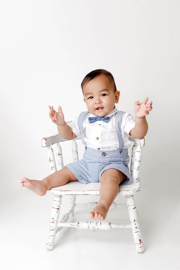 Adorable baby boy in blue suspenders and bow tie sitting in a white rustic chair by French Valley baby photographer.