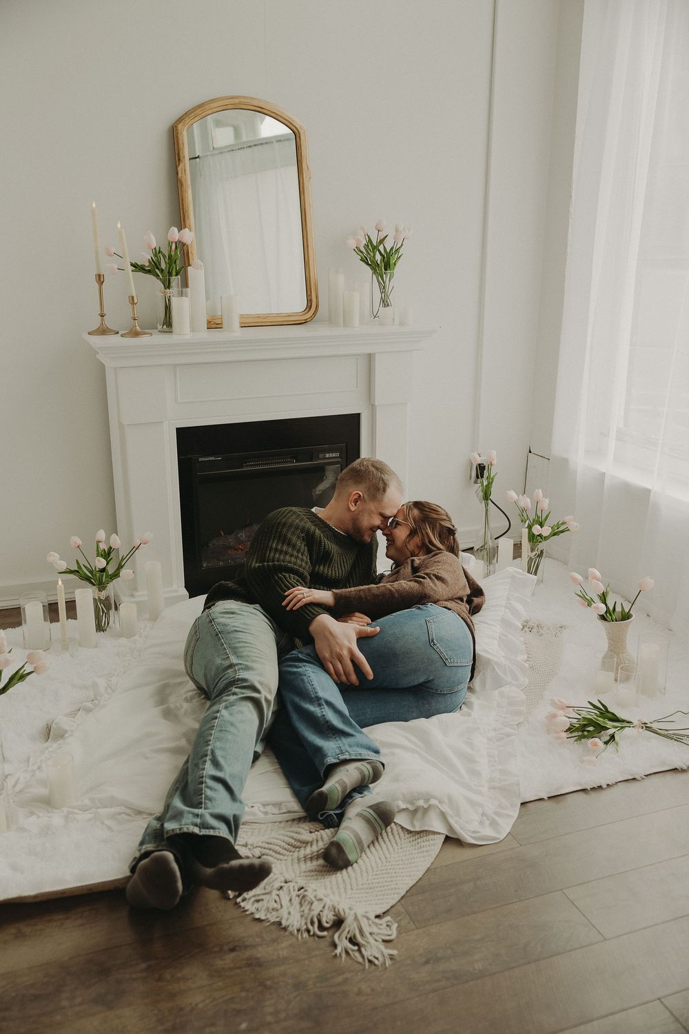 Romantic couple sitting together on a white blanket by a fireplace surrounded by pink tulips in a cozy living room.