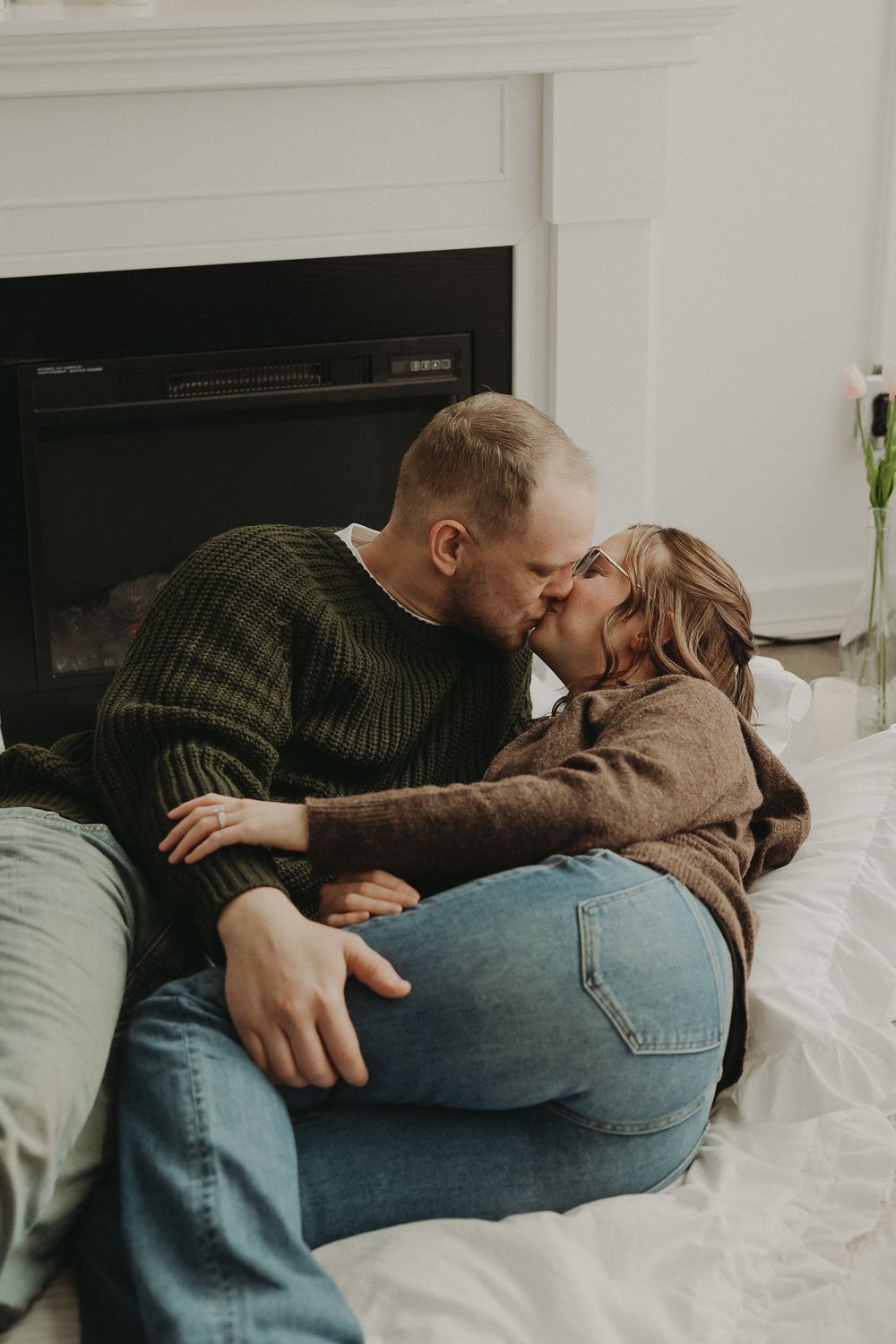 Couple sharing a romantic kiss by a fireplace, dressed in cozy sweaters and jeans, sitting on a white bed.