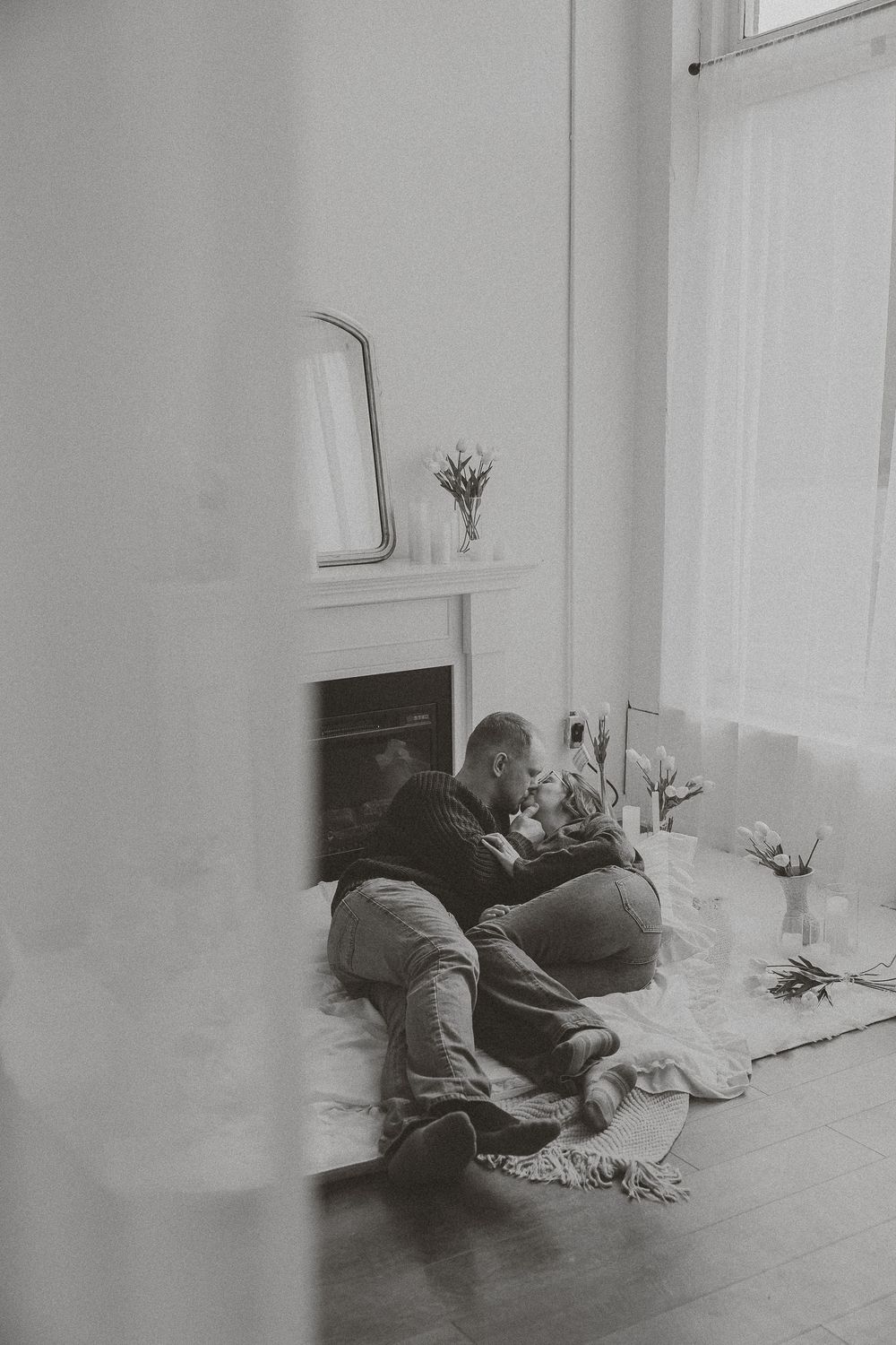 Black and white photo of a couple sitting together on the floor by a fireplace, sharing an intimate moment.