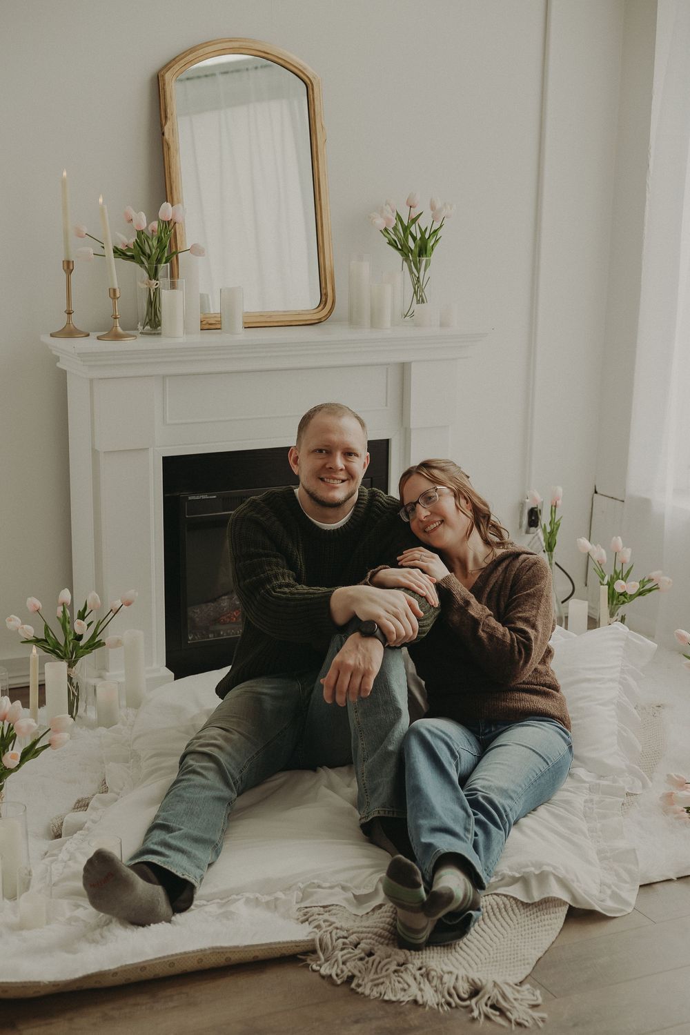 Happy couple sitting together on a blanket in front of a white fireplace decorated with candles and pink tulips.