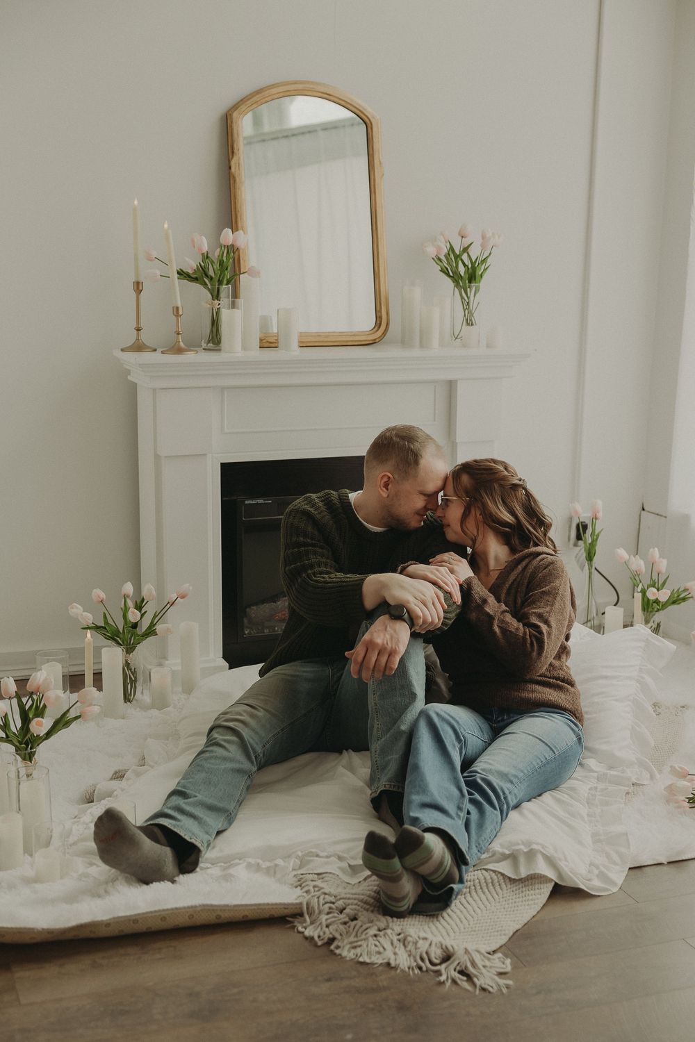 Romantic couple sitting together on a blanket by a white fireplace with tulips and candles in a cozy home setting.