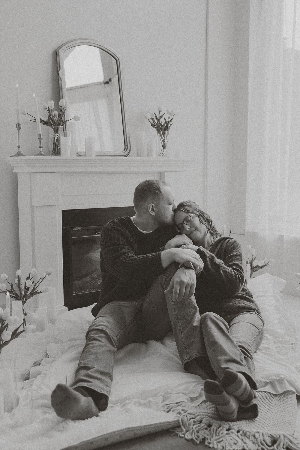 Black and white photo of a romantic couple sitting together on a blanket in front of a cozy fireplace with flowers nearby.