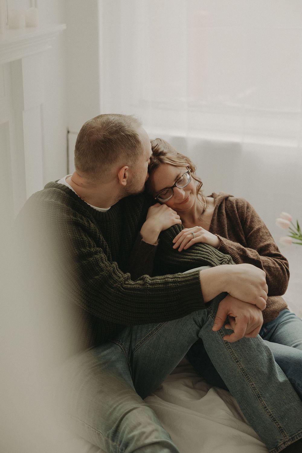 Couple sharing a tender moment, man kissing woman's cheek as she smiles, sitting together by a bright window.