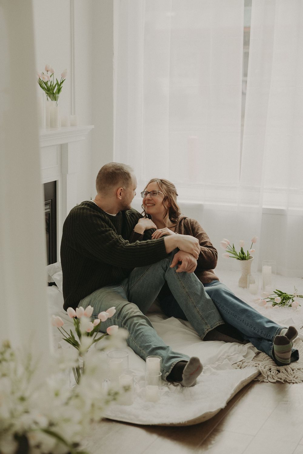 Couple sitting on floor by sheer curtains, surrounded by tulips, sharing a tender moment in a bright, cozy room.
