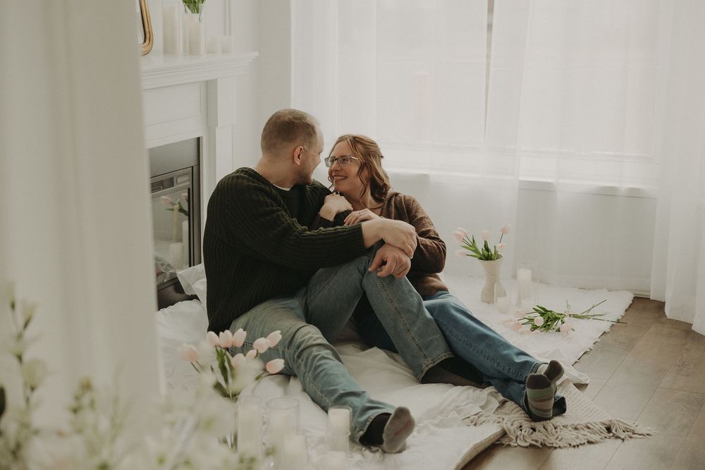 Couple sitting on floor sharing a tender moment surrounded by tulips in a bright, cozy white room.