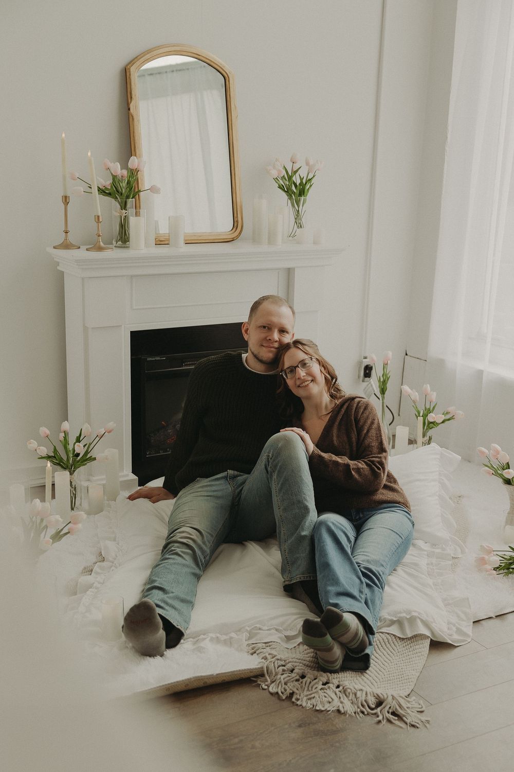 Couple sitting on white blanket by fireplace with pink tulips and candles in bright romantic room.