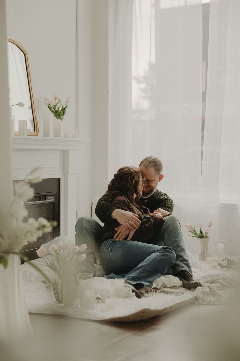 A couple sitting on the floor sharing a tender embrace near a white fireplace decorated with pink tulips.