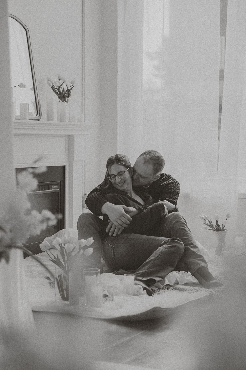 Black and white photo of a couple sitting on the floor, embracing and laughing near a fireplace with flowers.