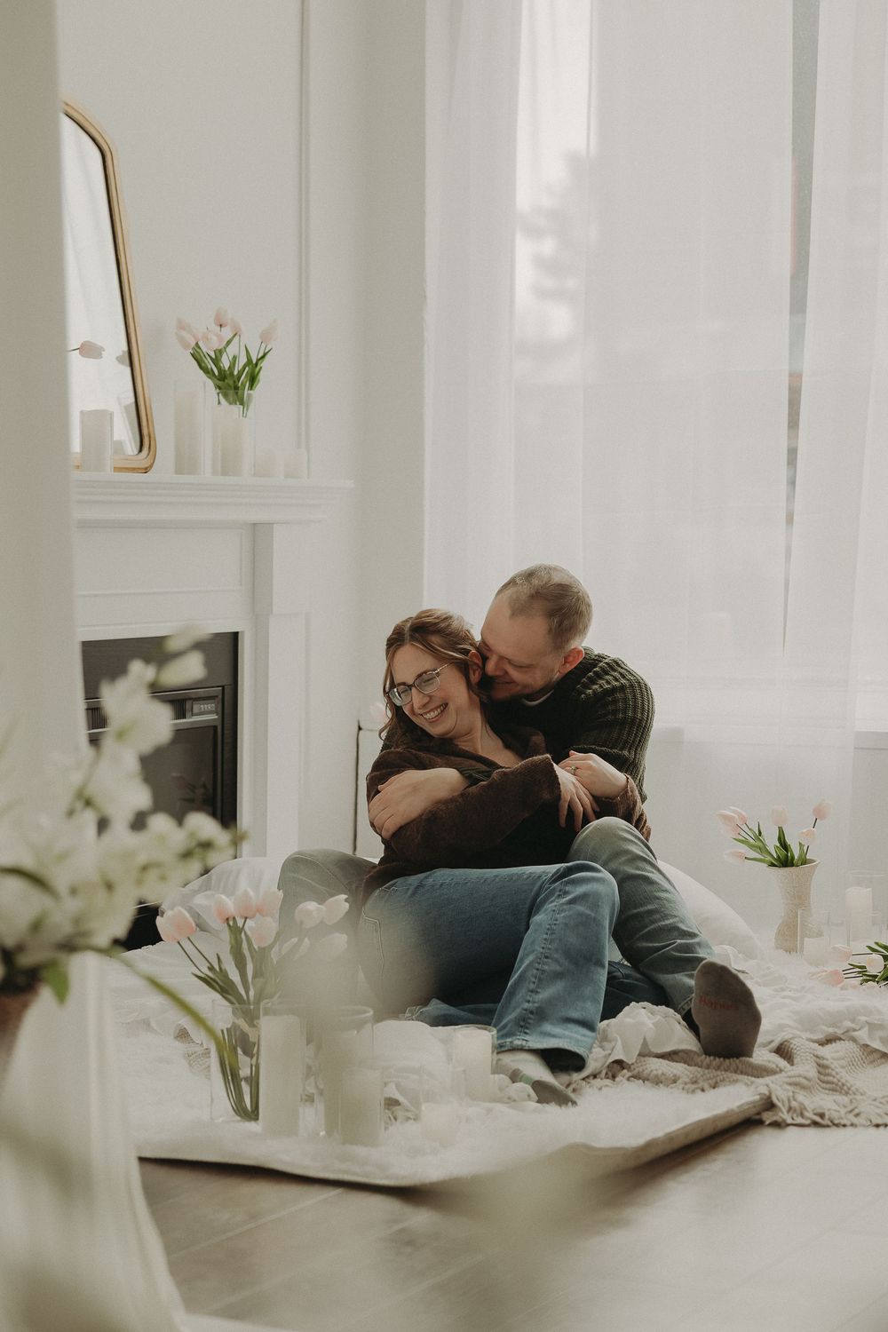 Couple embracing and laughing together on a blanket by a fireplace, surrounded by tulips in a bright white room.