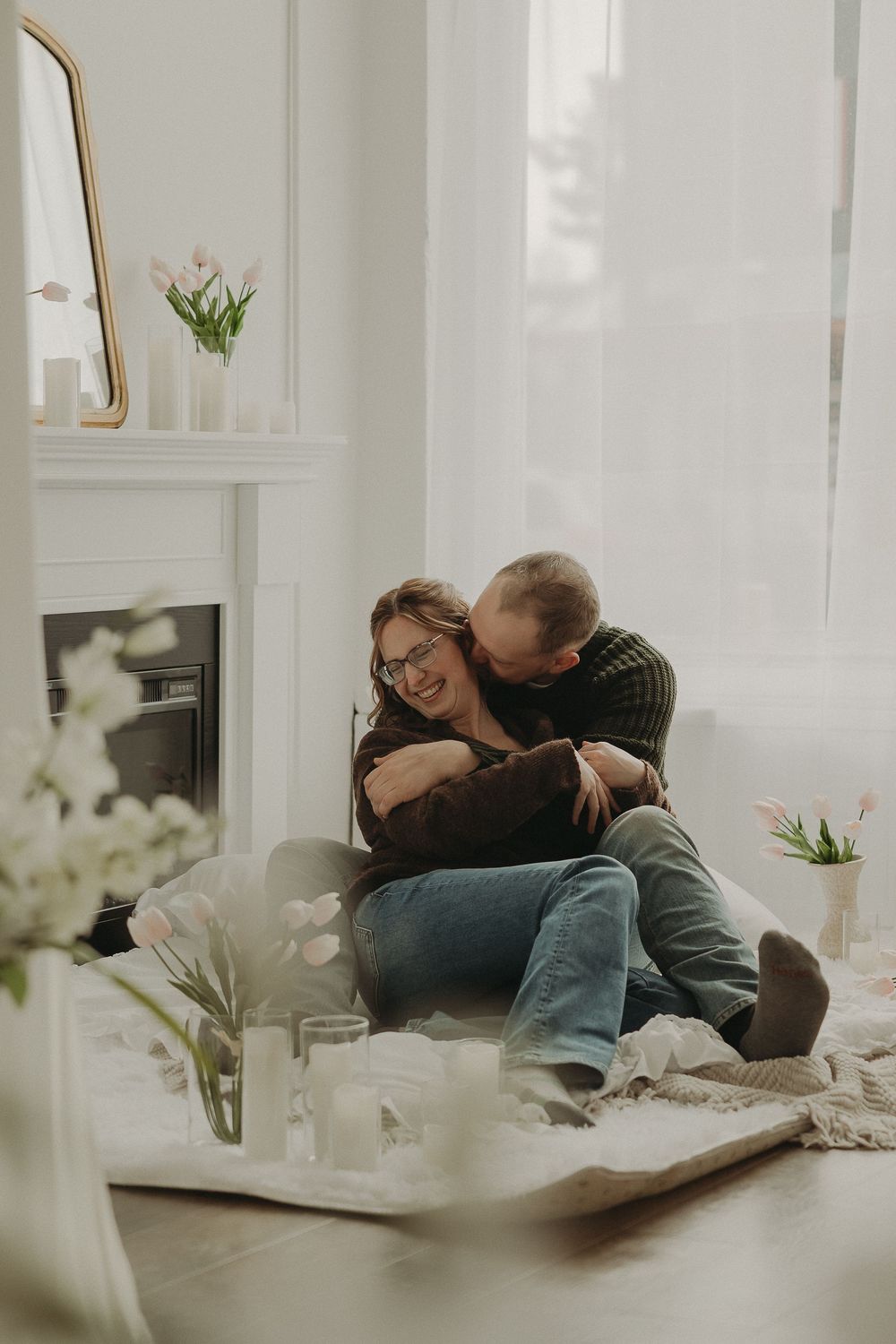 Couple embracing romantically on floor by fireplace in bright white living room with candles and fresh flowers.