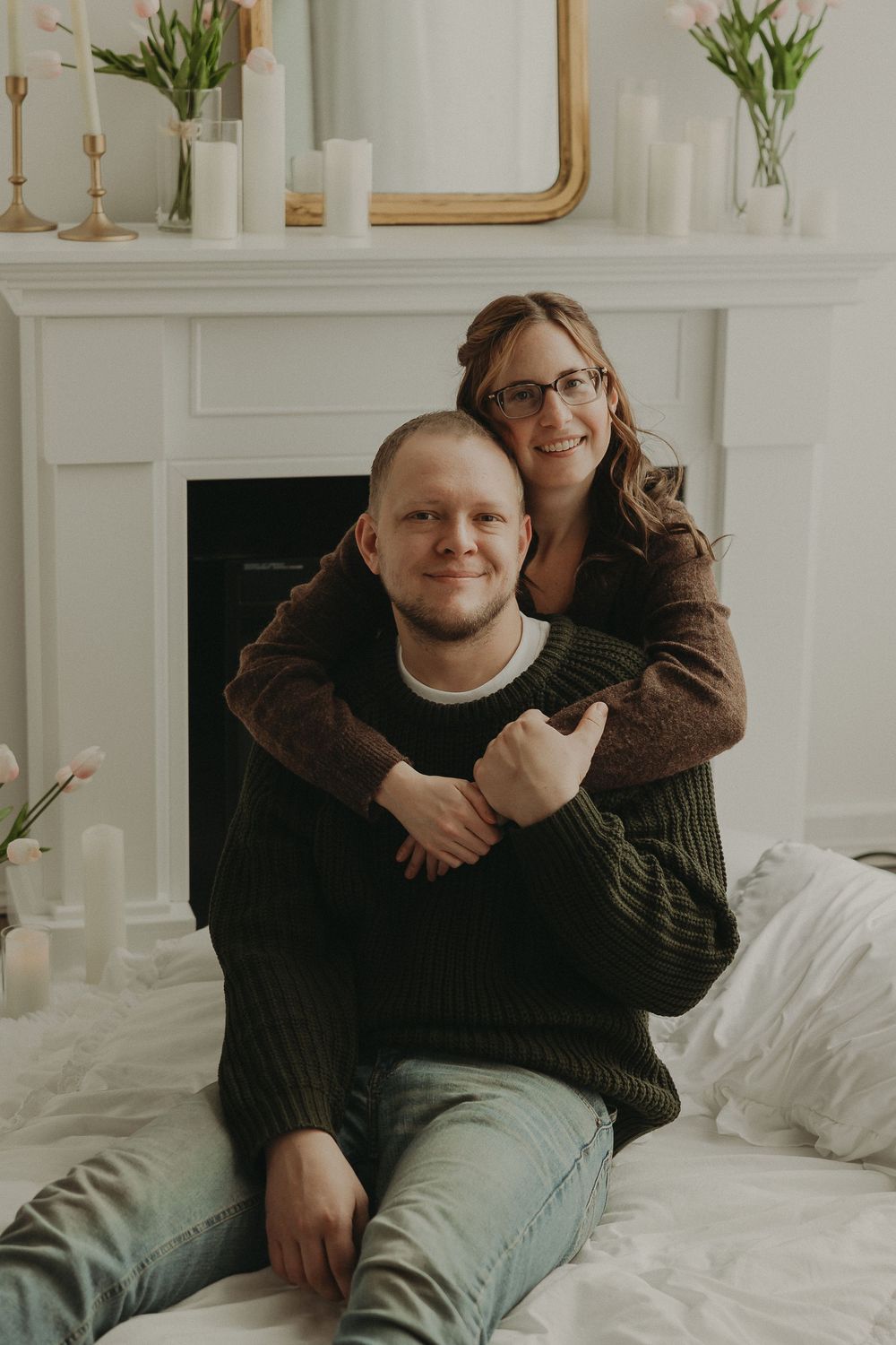 Couple embracing warmly in front of a white fireplace with candles and tulips, wearing cozy sweaters.