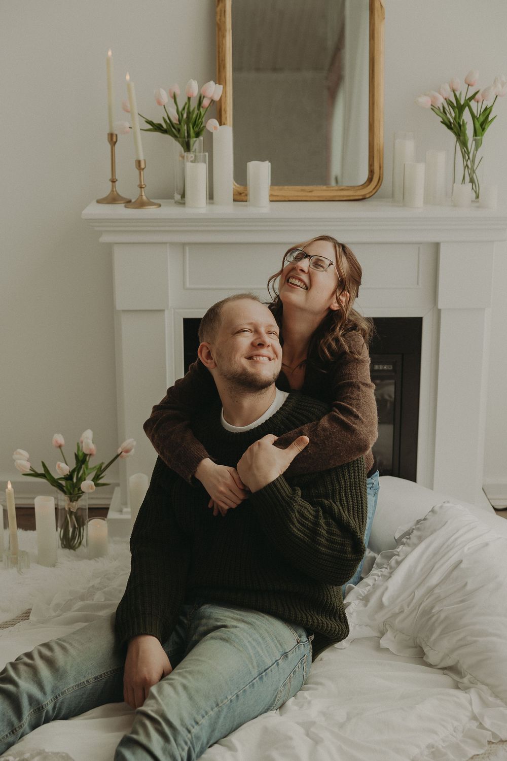 A smiling couple embracing near a white fireplace decorated with pink tulips, candles, and a gold mirror.