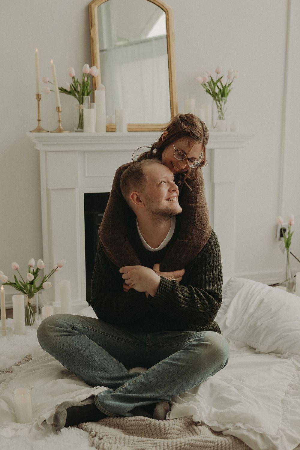 Couple embracing on a bed near a white fireplace decorated with pink tulips and candles in a cozy bedroom.