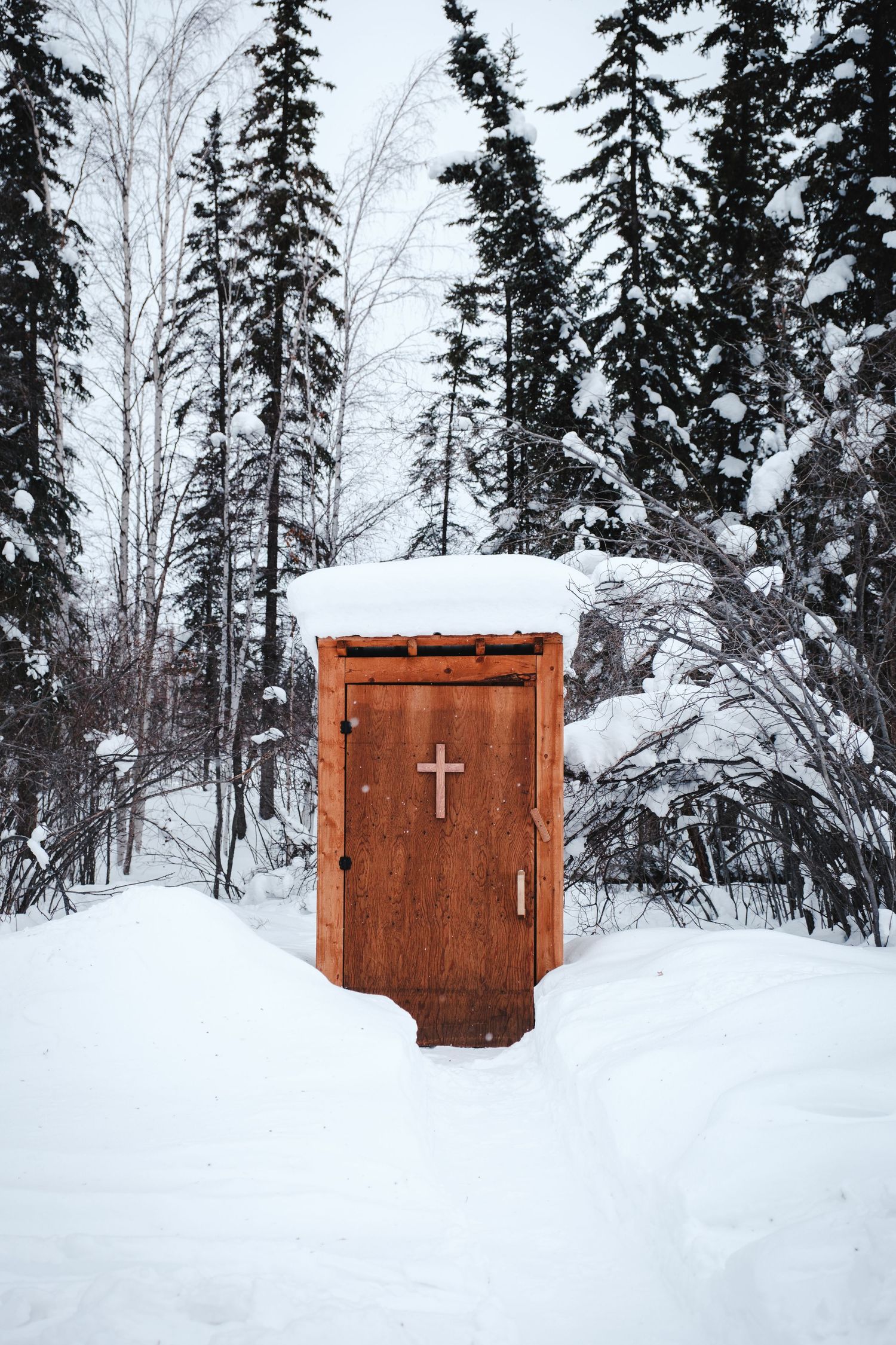 A rustic wooden outhouse with a cross on its door stands in a snowy winter forest with frost-covered trees.