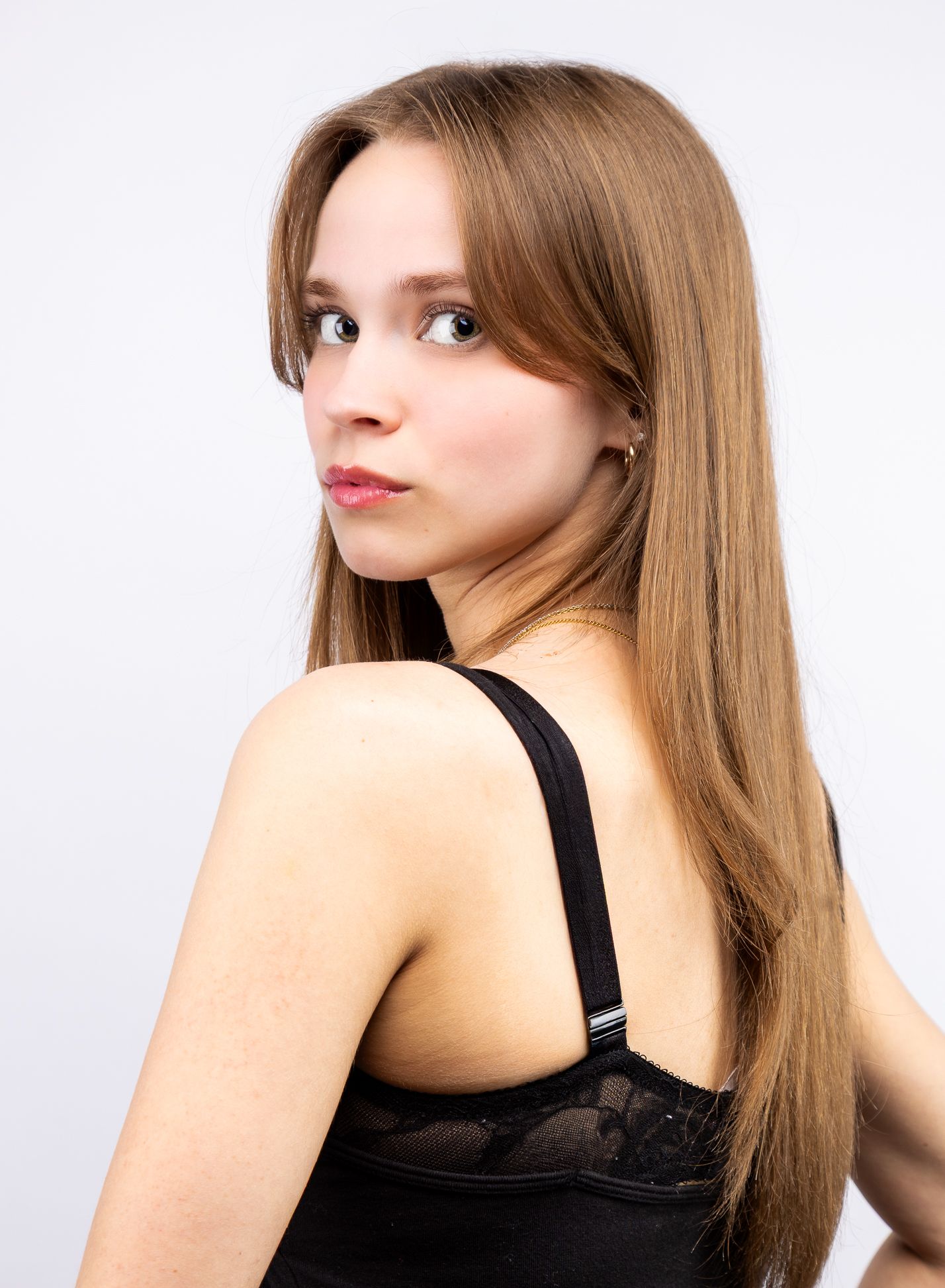 A girl in front of a white backdrop in a black tank top, looking over her shoulder to look at the camera