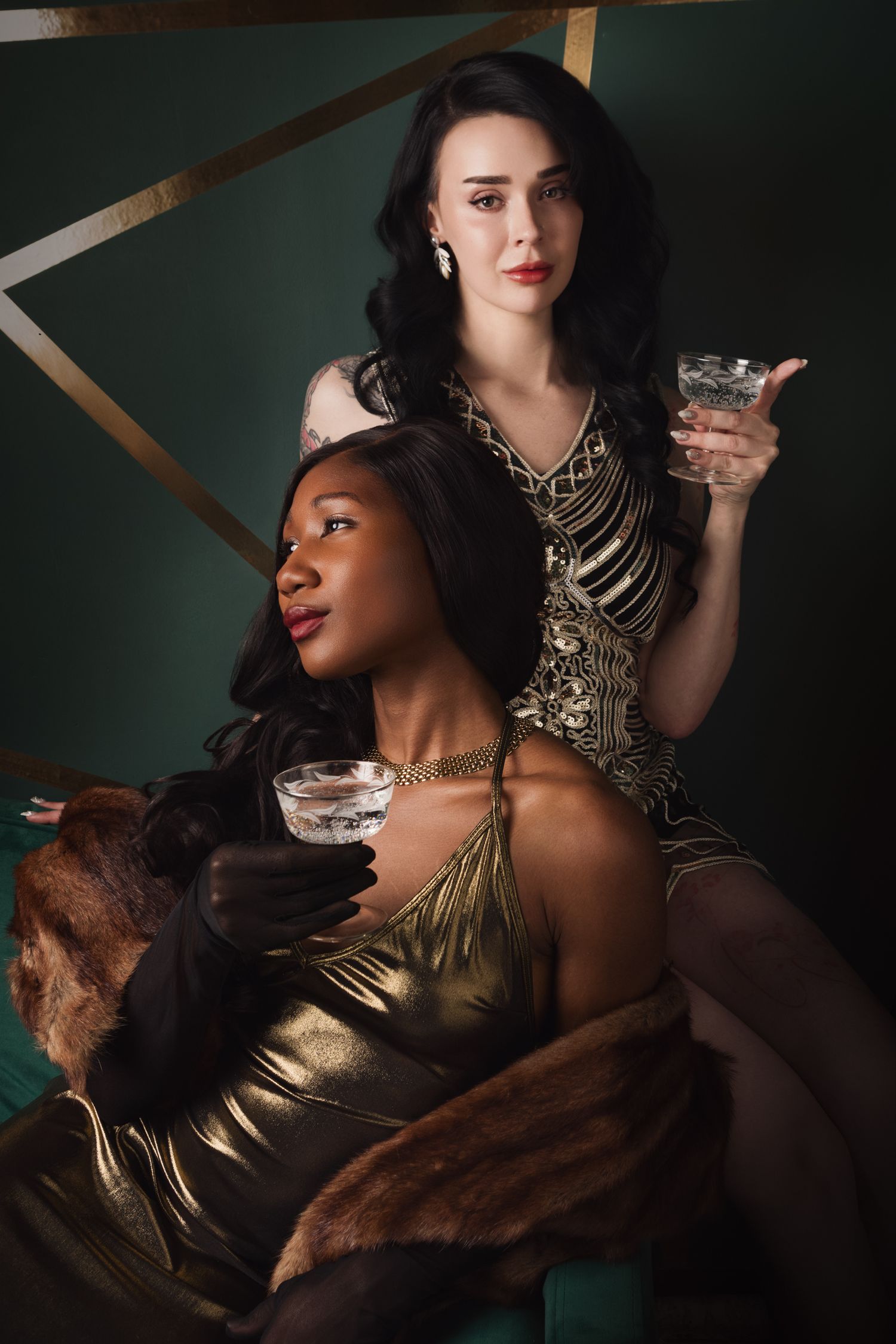 Two elegant women in gold dresses holding cocktail glasses pose glamorously against a dark green art deco background.