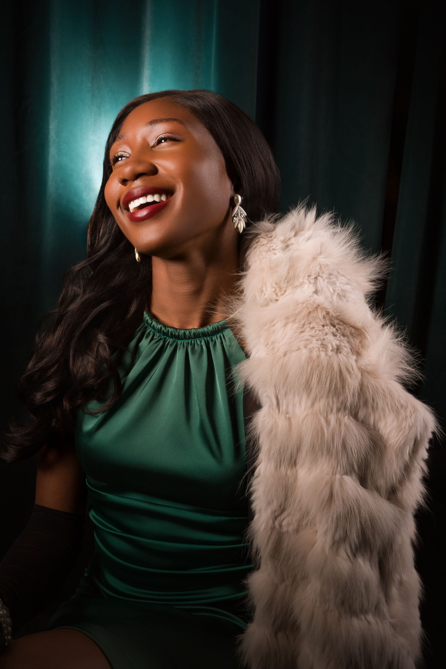 Elegant Black woman in emerald green satin halter gown with diamond earrings and black gloves, posed gracefully.