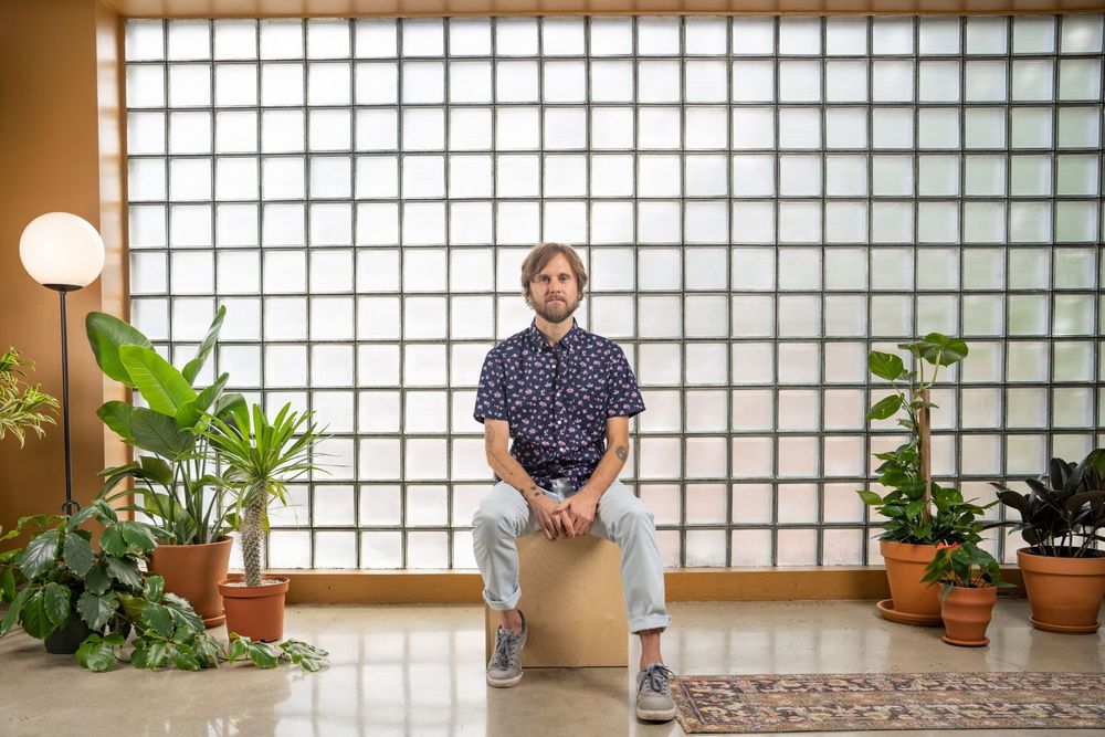 Wide editorial portrait of a man sitting in a bright, plant-filled studio with a gridded glass wall; environmental branding and professional