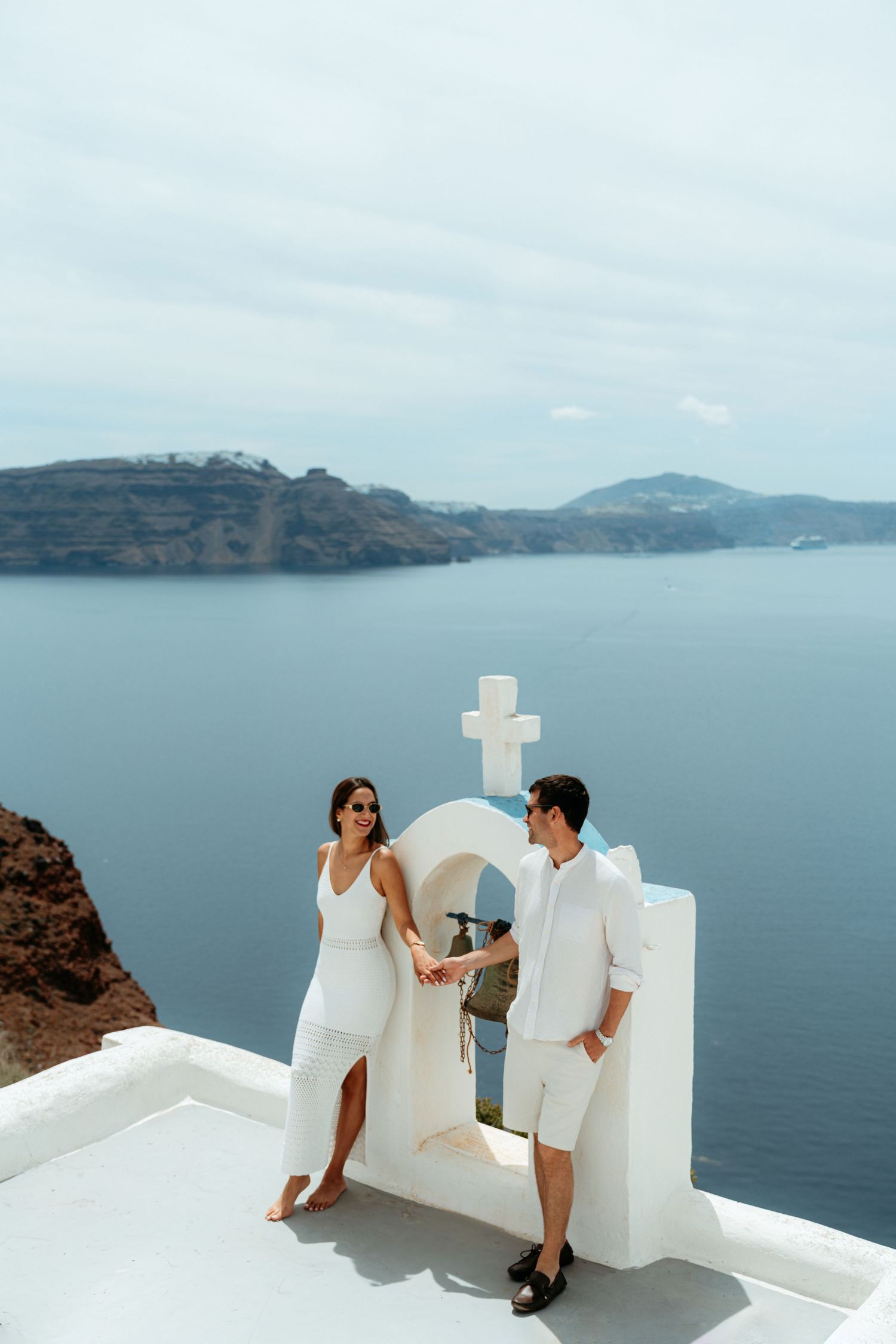 Couple in white outfits holding hands by a white arch with cross, overlooking Santorini caldera and sea.