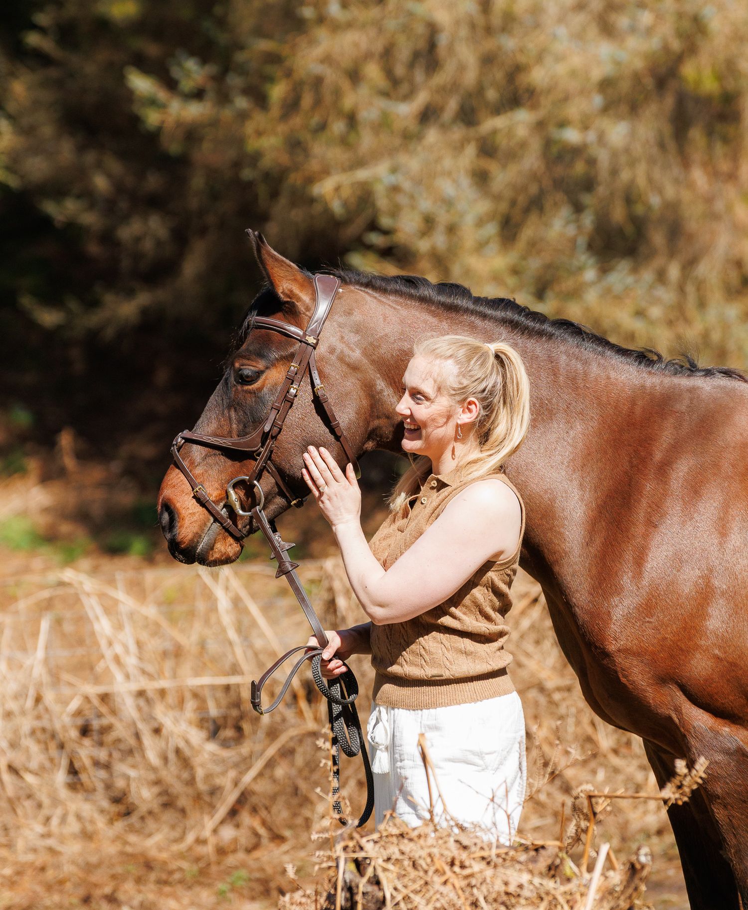 A smiling blonde woman in a brown vest affectionately touches a chestnut horse wearing a bridle outdoors.