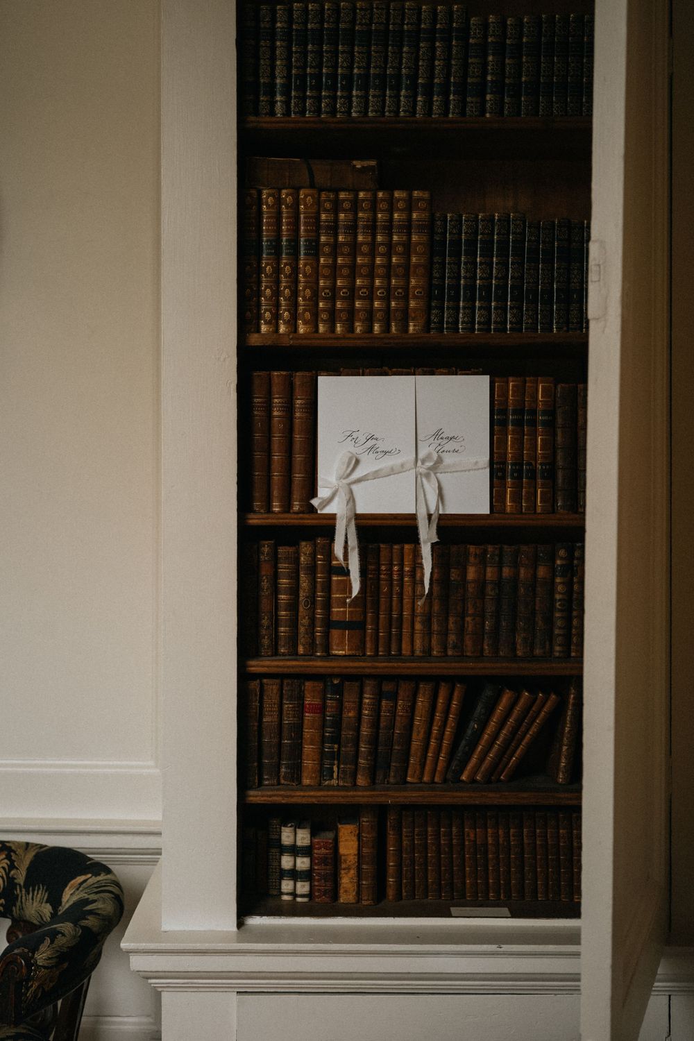 Elegant built-in wooden bookshelf filled with antique leather-bound books, featuring two white ribbon-tied wedding programs.