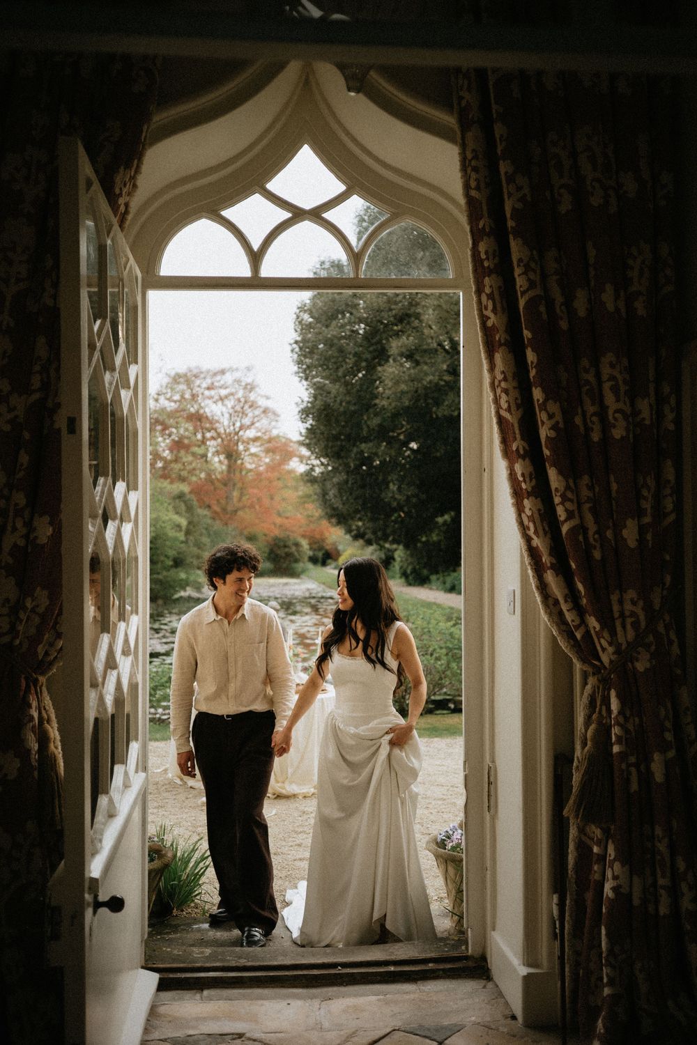 Bride and groom holding hands framed by ornate Gothic arch doorway with autumn garden views beyond.