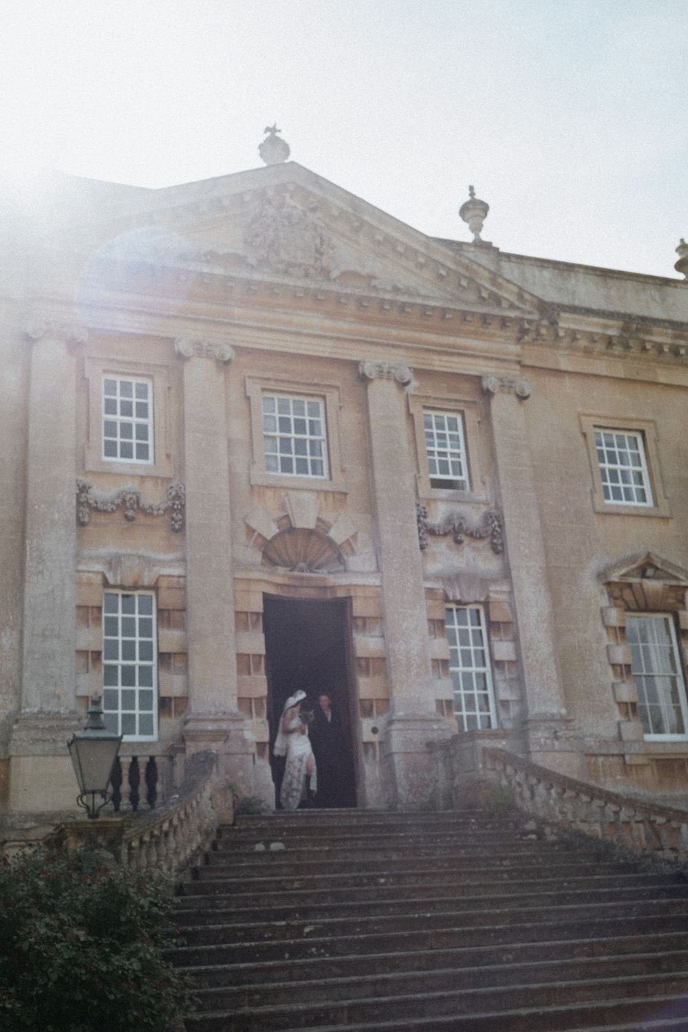 Couple embracing at entrance of grand Baroque stone mansion with tall columns and sweeping stone staircase.