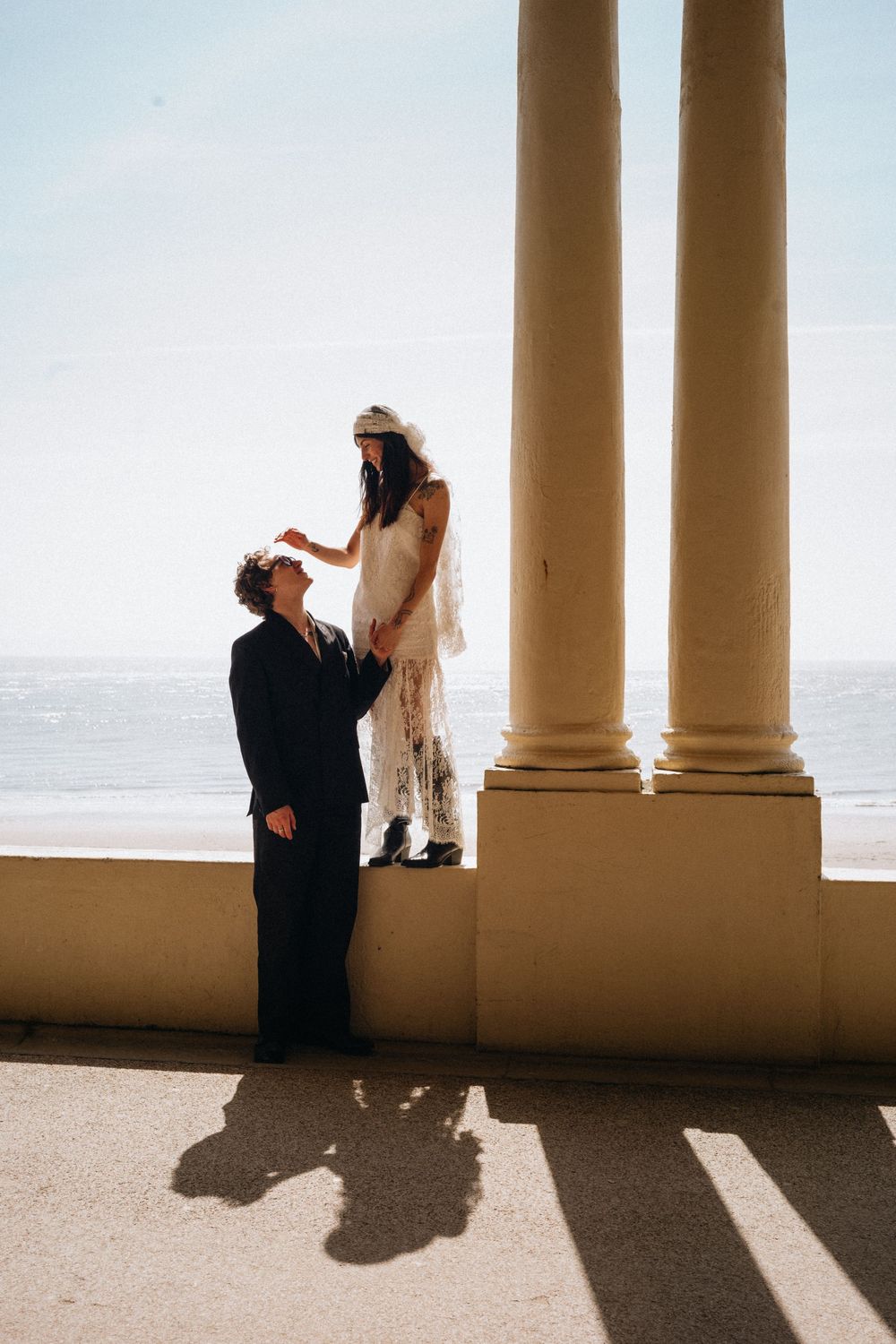 Bride standing on a balcony wall touching grooms face, with ocean views and classical columns in bright sunlight.