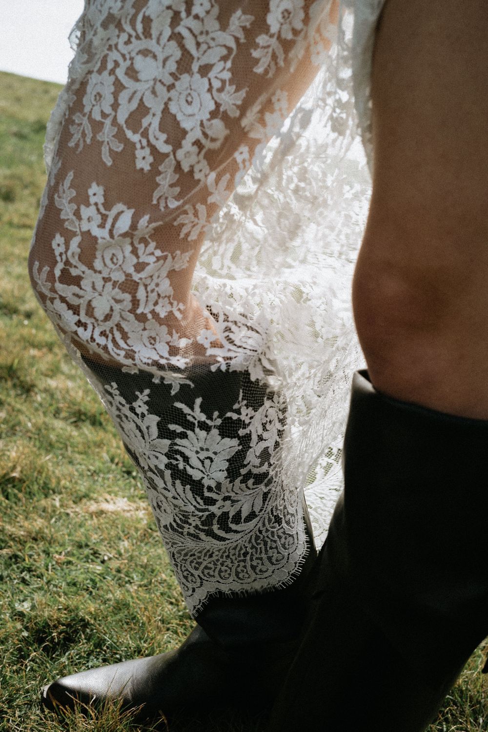 Close-up of a white lace wedding dress hem over black cowboy boots on green grass.