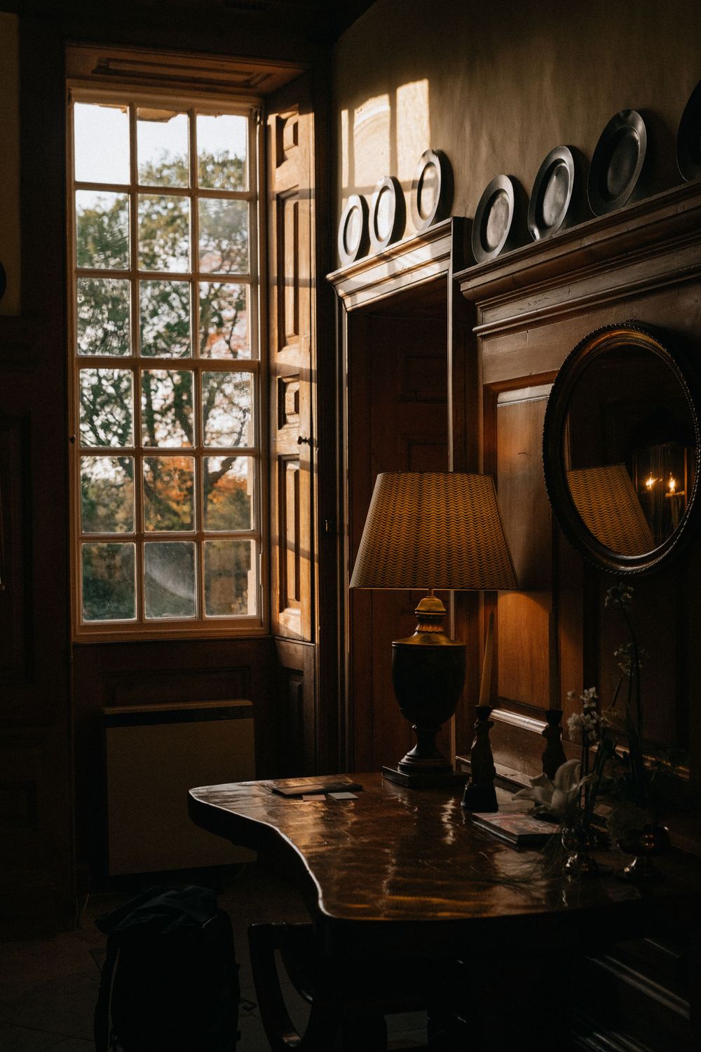 Warm golden sunlight streams through Georgian windows into a dark wood-panelled room with antique desk and brass lamp.