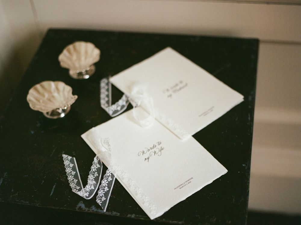Elegant wedding stationery with white cards, lace ribbon, and decorative shell dishes on a dark surface.