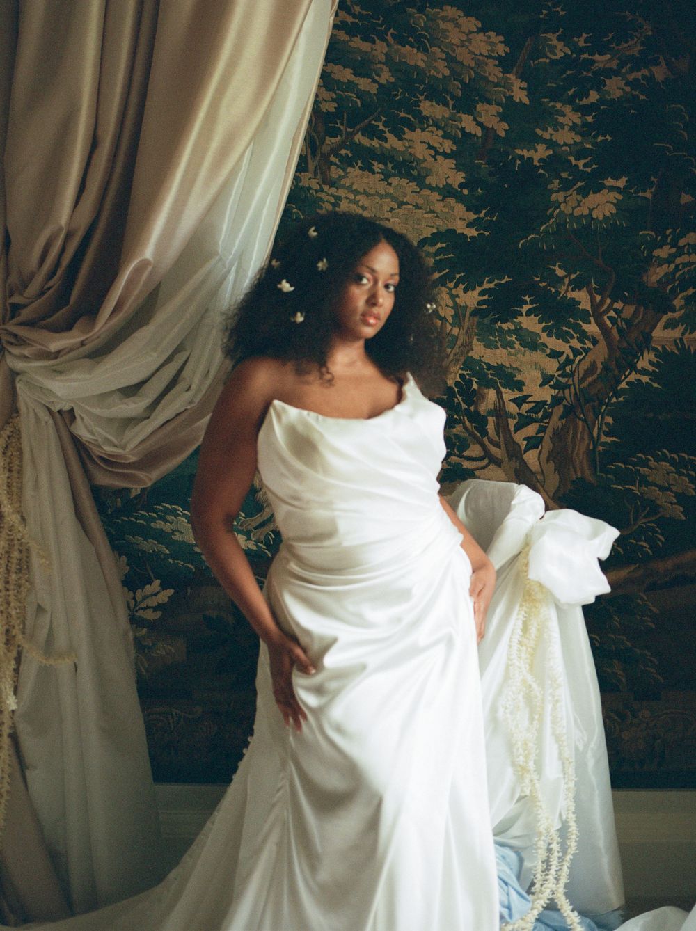 A Black woman in a strapless white satin wedding gown with bow detail stands before a tapestry backdrop.