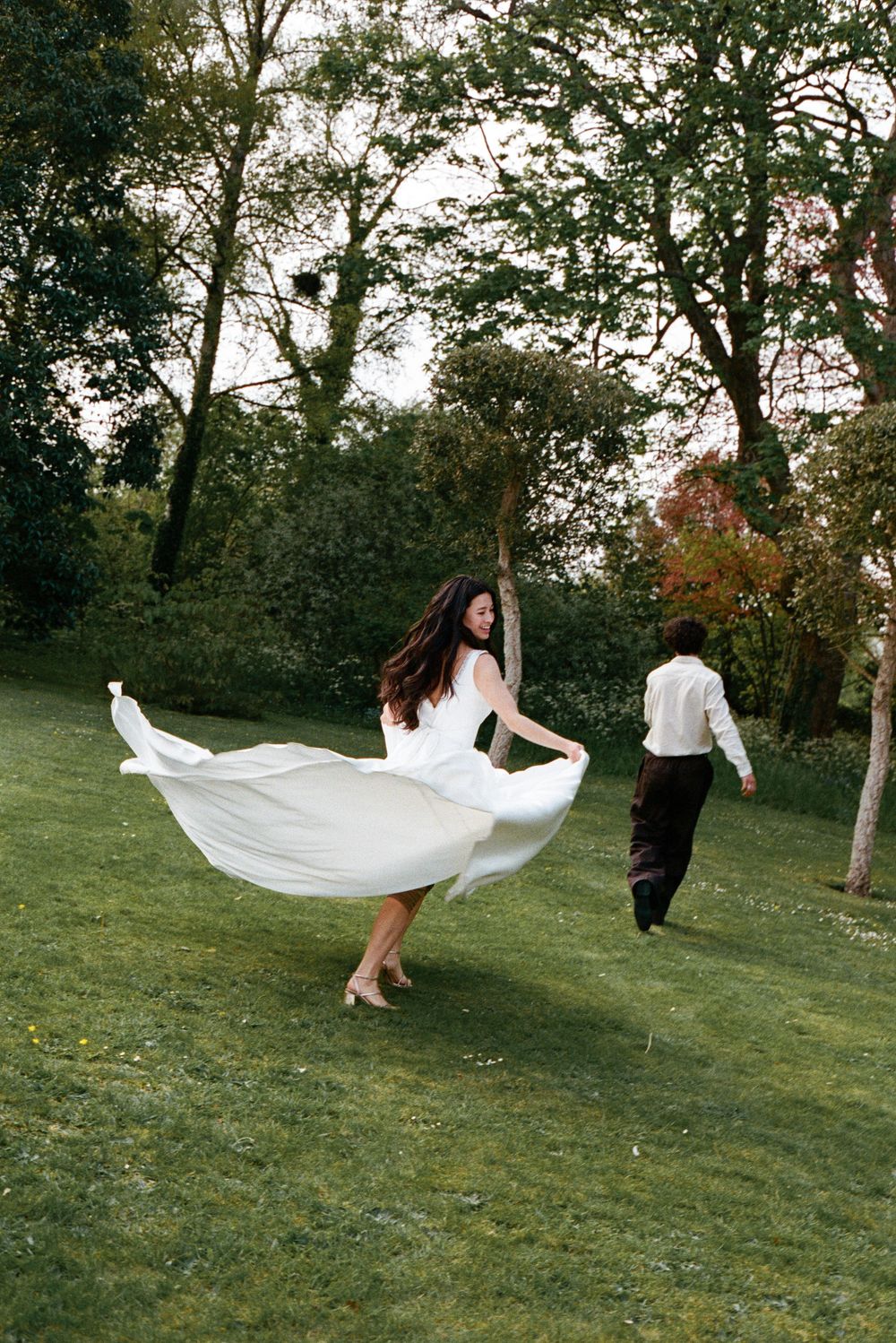 Bride twirls in flowing white wedding dress on lush green lawn as groom walks ahead among trees.