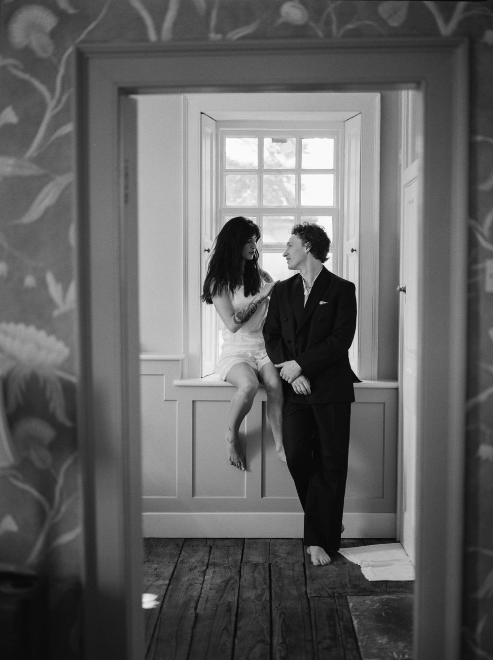 Black and white photo of a couple sharing a romantic moment by an open window, woman seated on sill, man standing close.