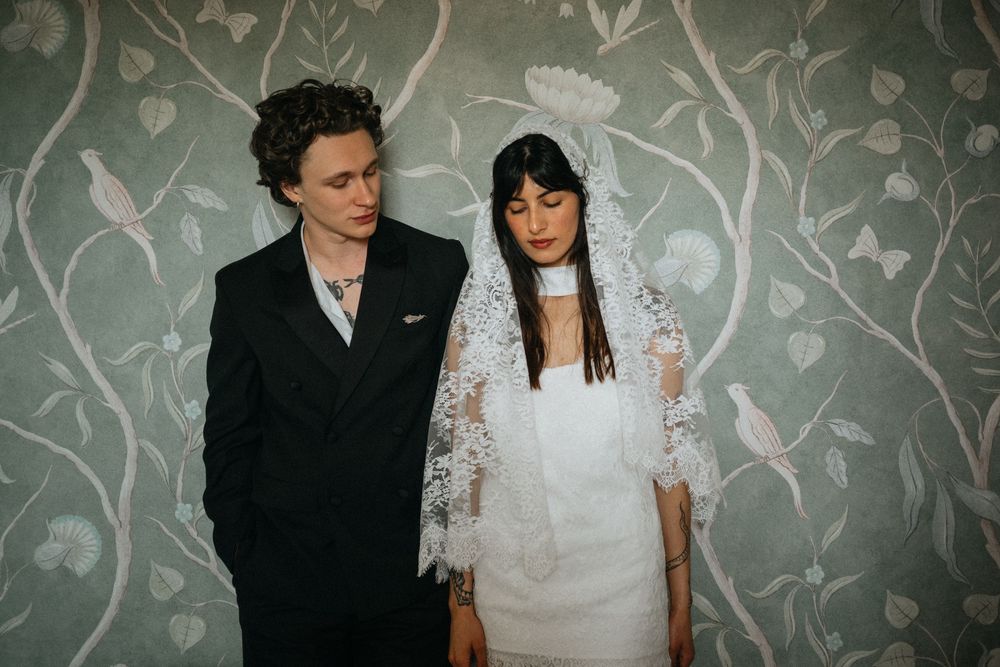 A groom in a black suit stands beside a bride wearing a white lace veil and dress against a floral wallpaper backdrop.