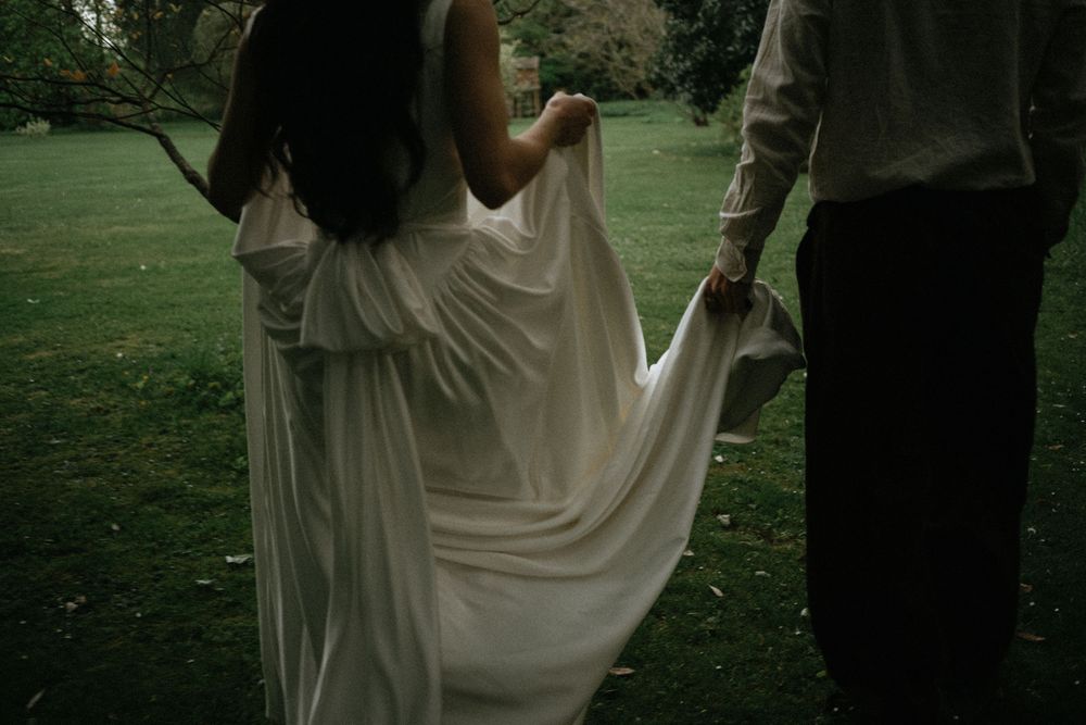 Bride in flowing white gown holds hands with groom outdoors on lush green grass in romantic wedding photo.