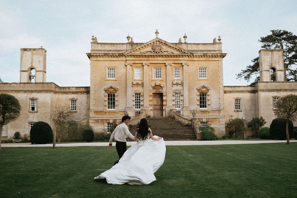 Couple dancing in front of a grand English country house mansion, bride in flowing white gown, lush green lawn.