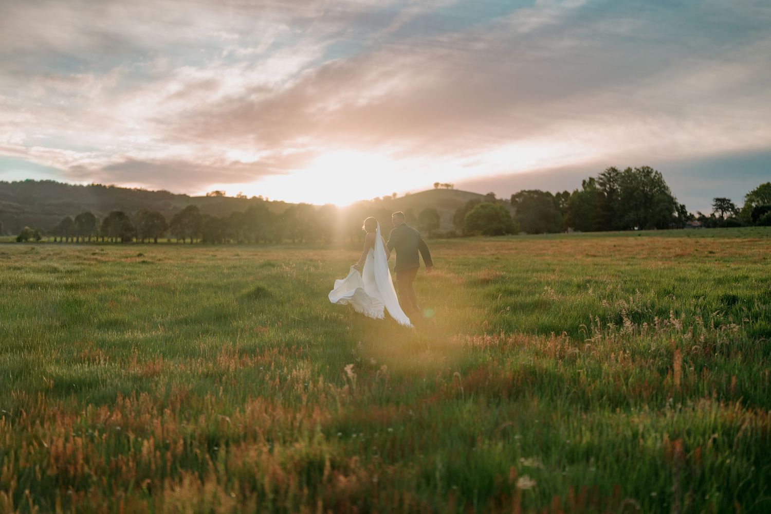A bride in a white dress stands in a lush green meadow at golden sunset, surrounded by wildflowers.