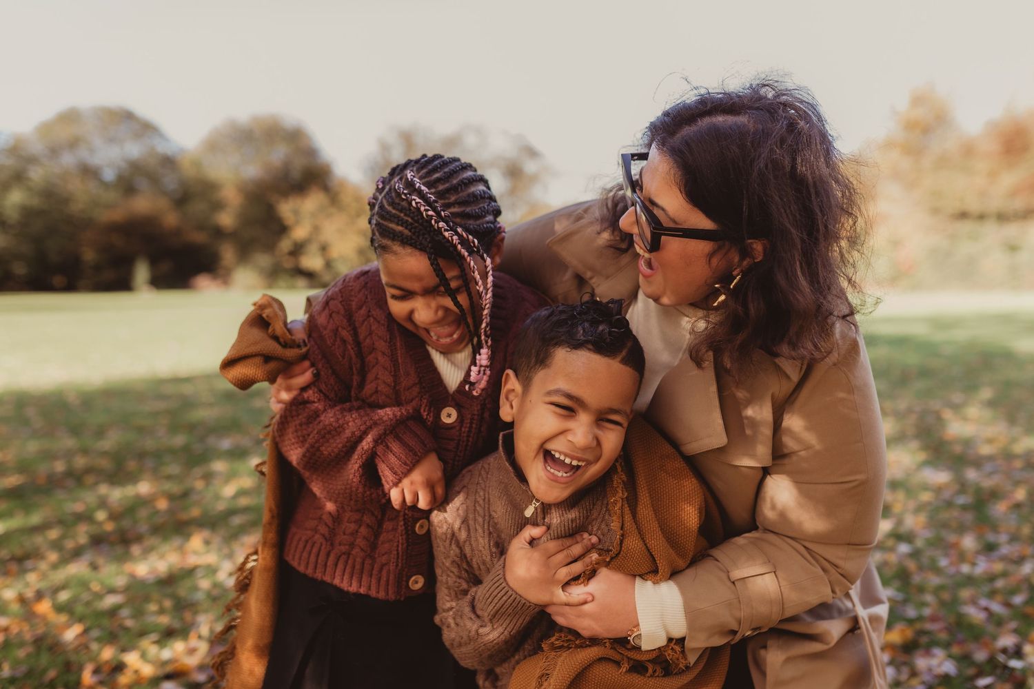 A mother hugging her two laughing children outdoors in a sunny autumn park setting.