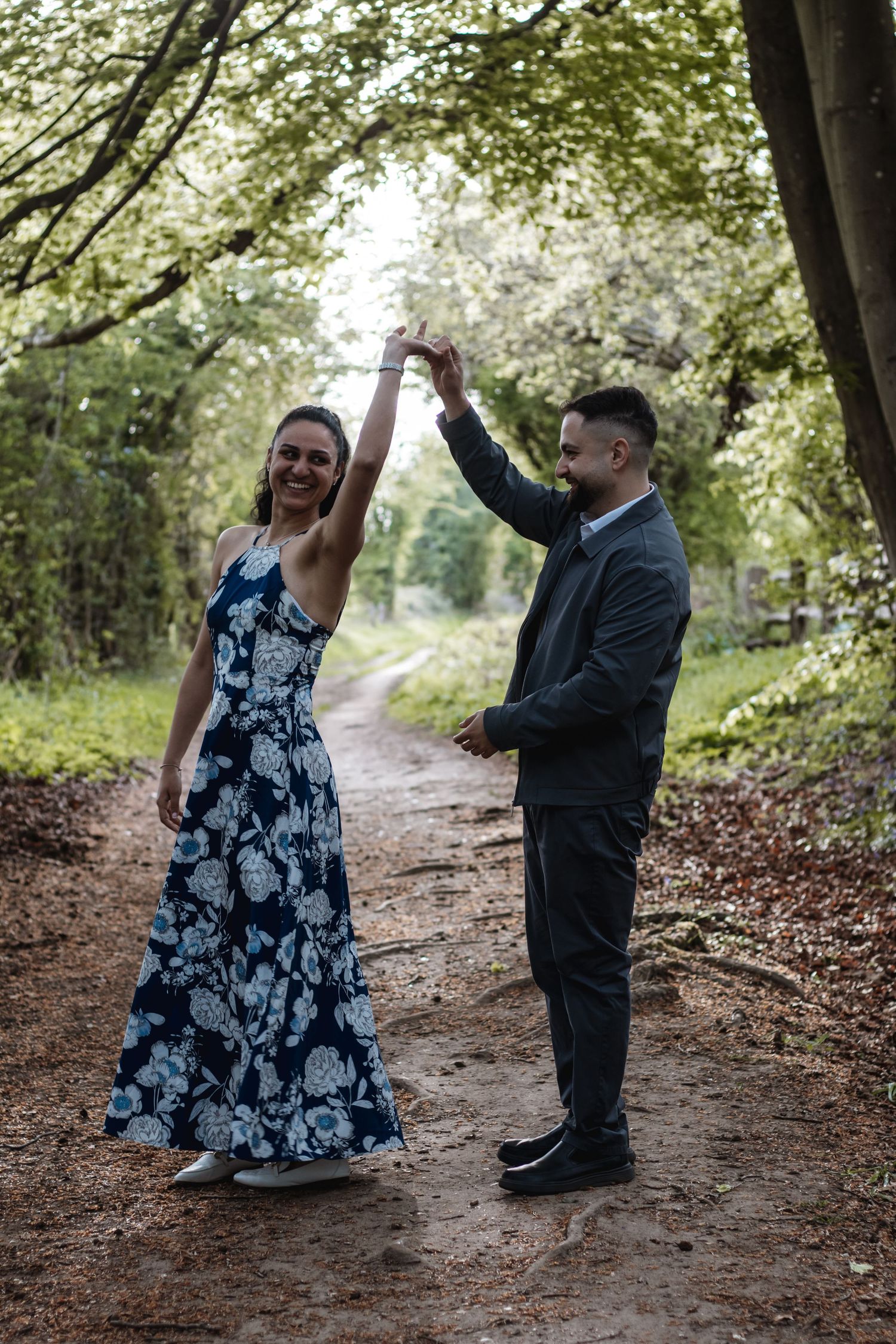 Couple dancing on a forest path, woman in floral dress twirled by man in gray suit.